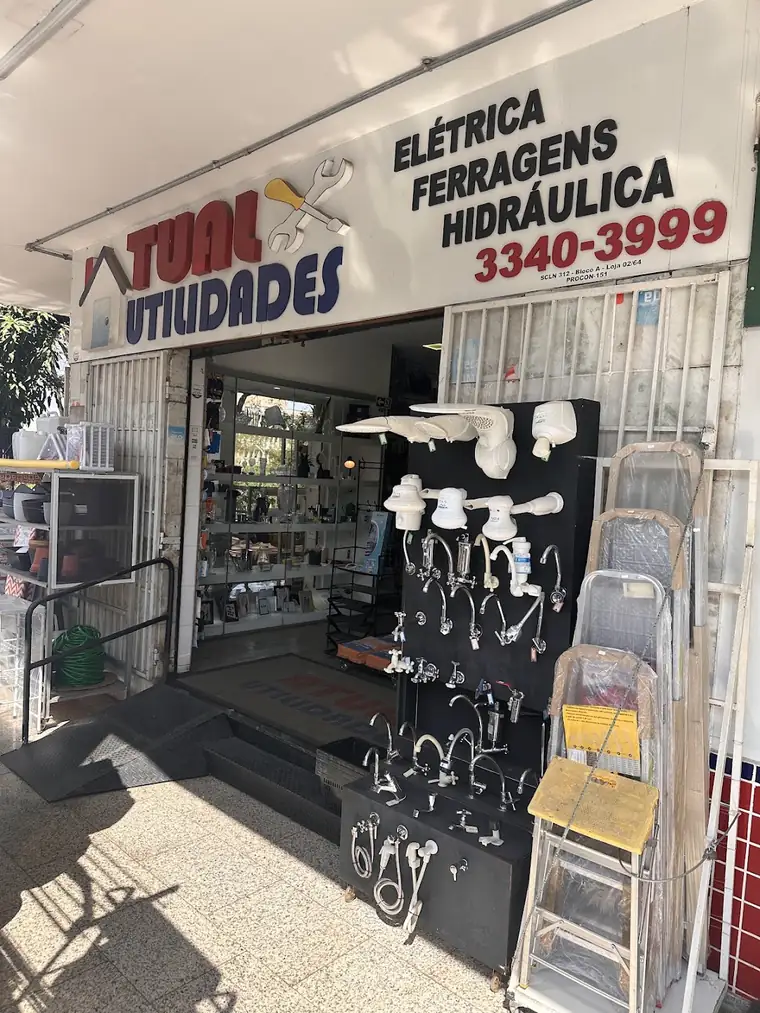 Fachada da Atual Utilidades: placa branca com ATUAL em vermelho, UTILIDADES em azul e as palavras elétrica, ferragens e hidráulica; na entrada, chuveiros, torneiras e escadas expostos